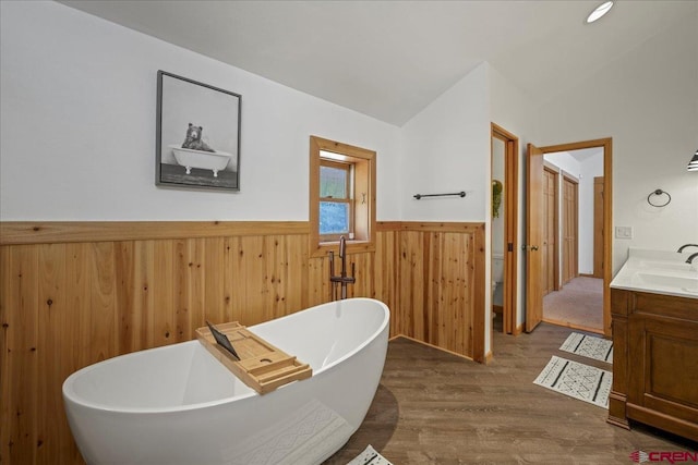 full bathroom featuring wood finished floors, vanity, a freestanding bath, vaulted ceiling, and wainscoting