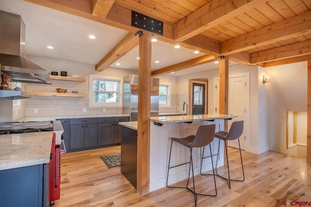 kitchen featuring light stone counters, light wood-style floors, high end fridge, wall chimney range hood, and beamed ceiling