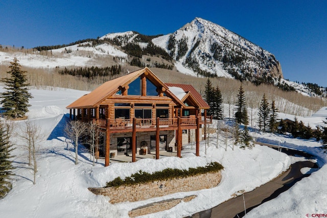 snow covered house featuring a deck with mountain view, stone siding, a patio area, and log exterior