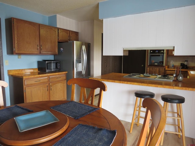 kitchen with a textured ceiling, appliances with stainless steel finishes, brown cabinetry, and a kitchen breakfast bar