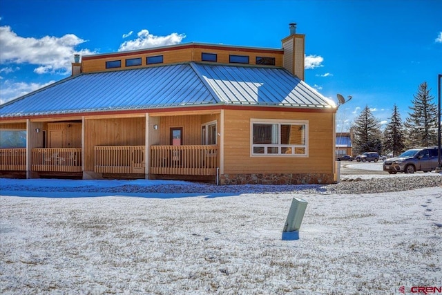 snow covered rear of property featuring a standing seam roof, covered porch, metal roof, and a chimney