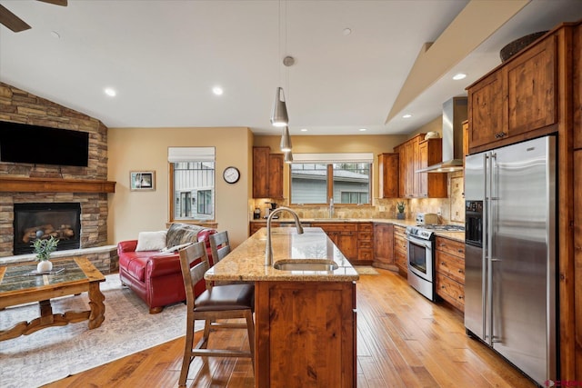 kitchen with appliances with stainless steel finishes, open floor plan, a sink, a stone fireplace, and wall chimney exhaust hood