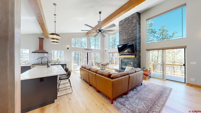 living area featuring beam ceiling, light wood finished floors, a high ceiling, ceiling fan, and a stone fireplace