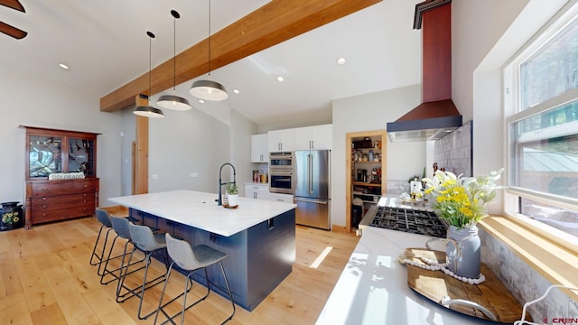 kitchen featuring wall chimney exhaust hood, vaulted ceiling with beams, stainless steel appliances, light wood-type flooring, and white cabinetry