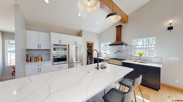 kitchen featuring light stone counters, light wood finished floors, stainless steel appliances, beamed ceiling, and wall chimney exhaust hood