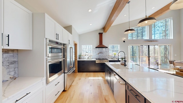 kitchen with light wood finished floors, appliances with stainless steel finishes, beamed ceiling, wall chimney range hood, and a sink