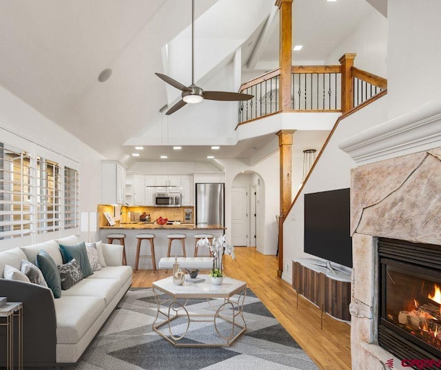 living area with arched walkways, a ceiling fan, a glass covered fireplace, high vaulted ceiling, and light wood-type flooring