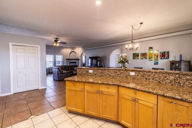 kitchen with light tile patterned floors, ceiling fan with notable chandelier, a fireplace, hanging light fixtures, and dark stone counters