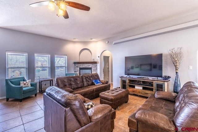 living room featuring arched walkways, a textured ceiling, a stone fireplace, light tile patterned flooring, and a ceiling fan