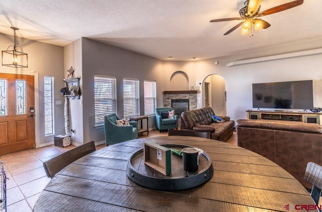 tiled dining room with arched walkways, ceiling fan, a stone fireplace, a textured ceiling, and baseboards