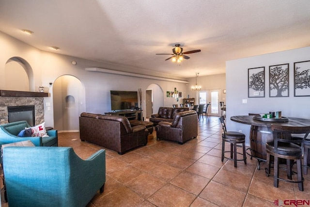 living room featuring arched walkways, tile patterned flooring, a stone fireplace, and ceiling fan with notable chandelier