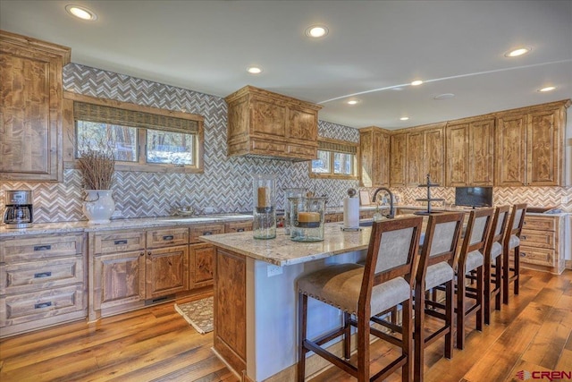 kitchen with brown cabinets, light wood finished floors, a kitchen island with sink, light stone countertops, and a kitchen breakfast bar