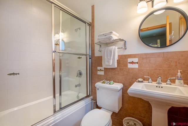 bathroom featuring tile walls, wainscoting, toilet, enclosed tub / shower combo, and a sink