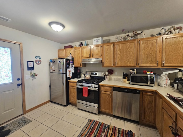 kitchen featuring light tile patterned floors, light countertops, appliances with stainless steel finishes, brown cabinetry, and under cabinet range hood