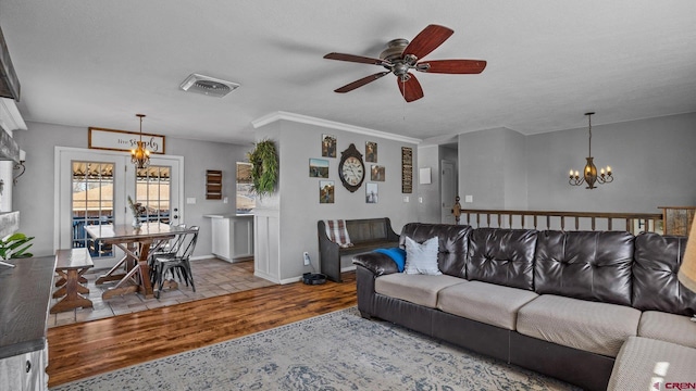 living area featuring wood finished floors, visible vents, baseboards, ornamental molding, and ceiling fan with notable chandelier