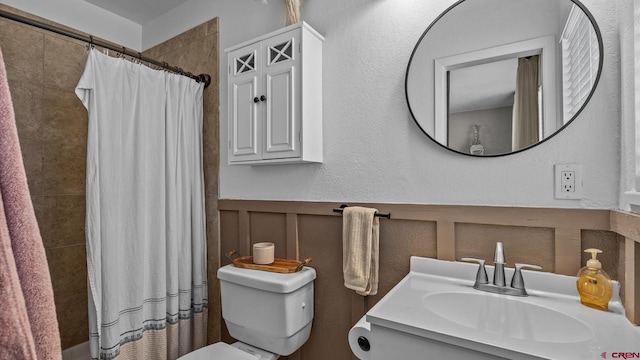 bathroom with a wainscoted wall, toilet, a tile shower, a decorative wall, and vanity