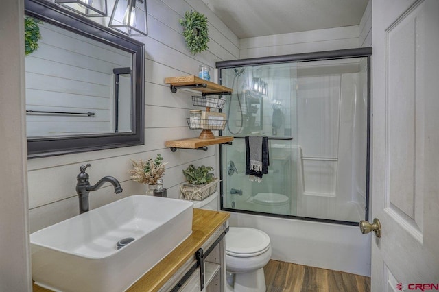 bathroom with vanity, wood finished floors, shower / bath combination with glass door, wood walls, and toilet