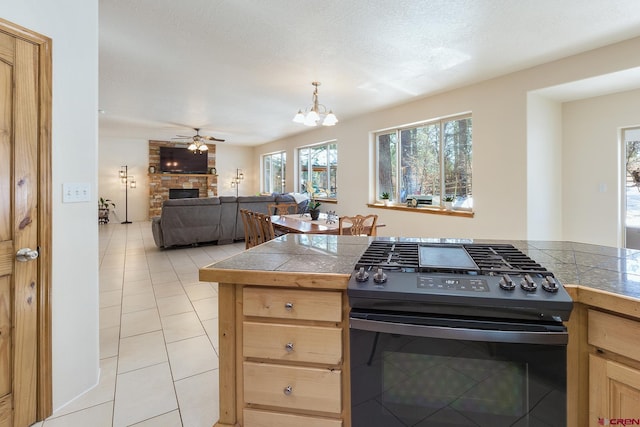 kitchen with light brown cabinetry, ceiling fan with notable chandelier, black gas stove, a stone fireplace, and light tile patterned flooring