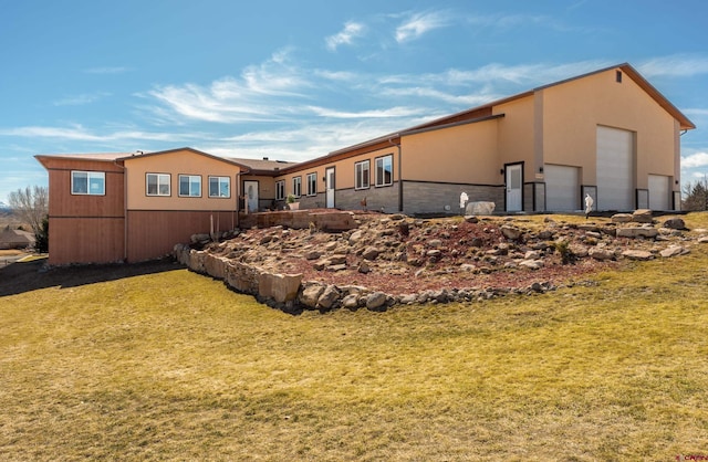 back of house with stucco siding, stone siding, a lawn, and an outbuilding