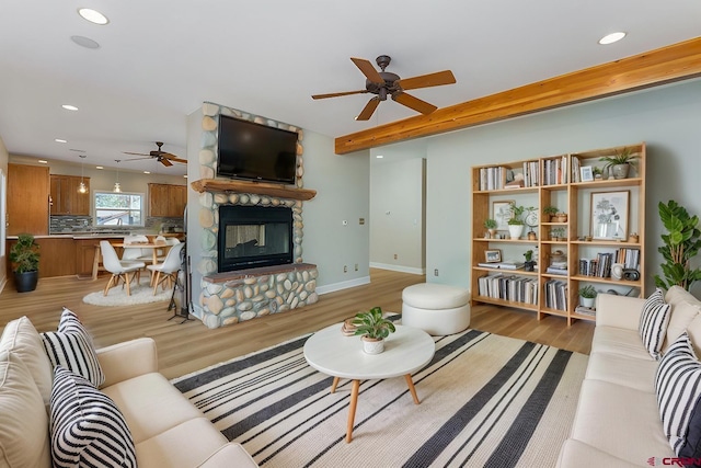 living area featuring baseboards, ceiling fan, beamed ceiling, light wood-type flooring, and a stone fireplace