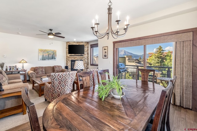 dining area featuring light wood-type flooring, a stone fireplace, and ceiling fan with notable chandelier