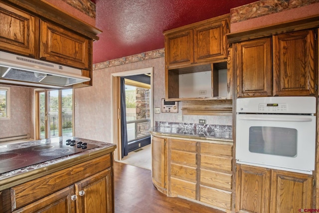 kitchen with black electric cooktop, wallpapered walls, dark wood-style floors, under cabinet range hood, and white oven