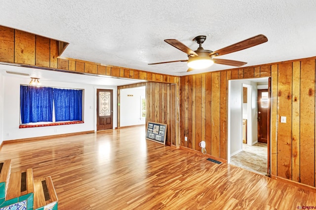 unfurnished living room featuring ceiling fan, a textured ceiling, wood finished floors, baseboards, and wooden walls