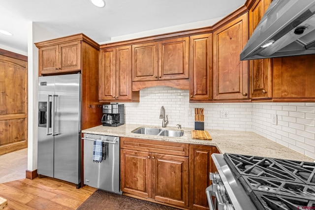 kitchen featuring brown cabinetry, under cabinet range hood, light stone counters, a sink, and stainless steel appliances