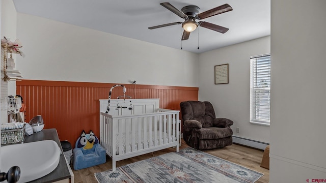 bedroom with a baseboard radiator, wood finished floors, a ceiling fan, a nursery area, and wainscoting