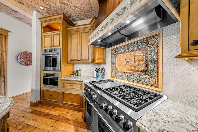 kitchen with wall chimney range hood, stainless steel appliances, backsplash, brick ceiling, and brown cabinetry