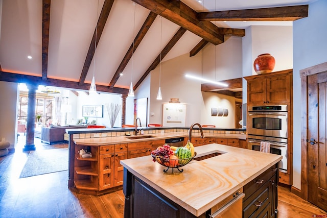 kitchen with a sink, stainless steel double oven, an island with sink, open floor plan, and beam ceiling