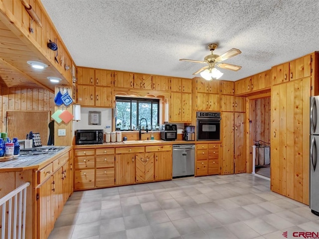 kitchen with black appliances, a sink, tile countertops, wood walls, and a ceiling fan
