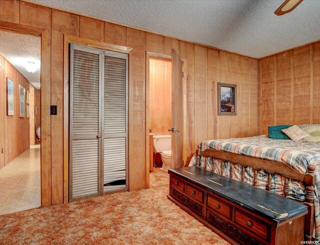 carpeted bedroom with a textured ceiling, a ceiling fan, a closet, wooden walls, and ensuite bath