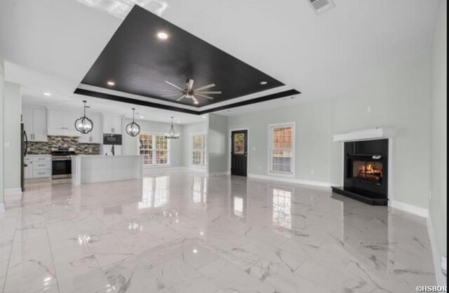 unfurnished living room featuring marble finish floor, a raised ceiling, a glass covered fireplace, baseboards, and ceiling fan with notable chandelier