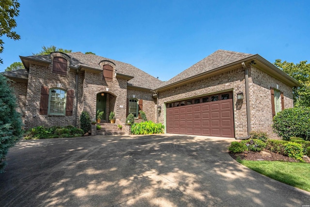 french provincial home with driveway, a shingled roof, a garage, and brick siding