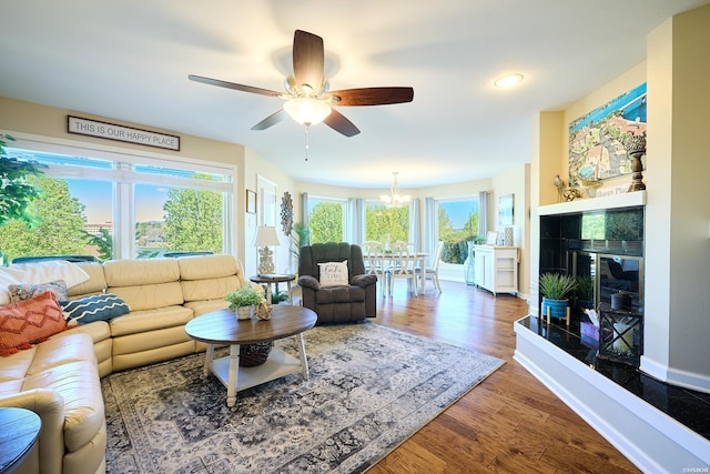 living area featuring ceiling fan with notable chandelier, a fireplace, dark wood finished floors, and baseboards