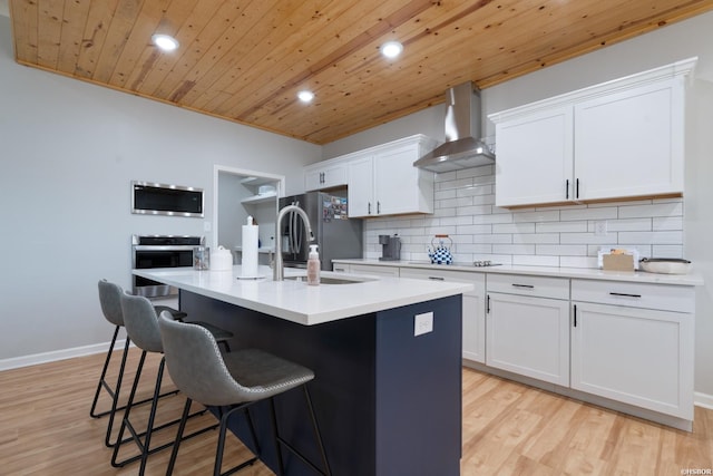 kitchen with light countertops, appliances with stainless steel finishes, a kitchen island with sink, white cabinetry, and wall chimney exhaust hood