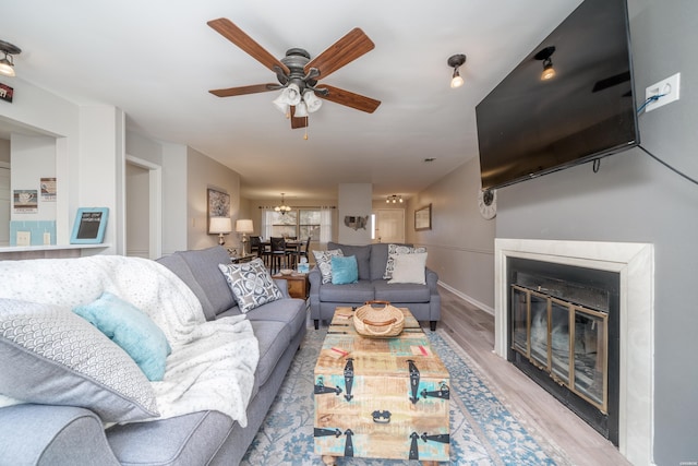 living area with light wood-type flooring, a glass covered fireplace, baseboards, and ceiling fan with notable chandelier