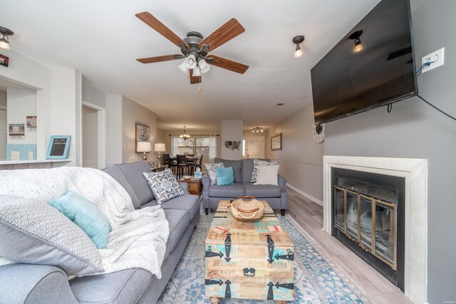 living area with light wood-type flooring, a glass covered fireplace, baseboards, and ceiling fan with notable chandelier