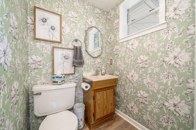 bathroom featuring vanity, toilet, baseboards, wallpapered walls, and wood finished floors