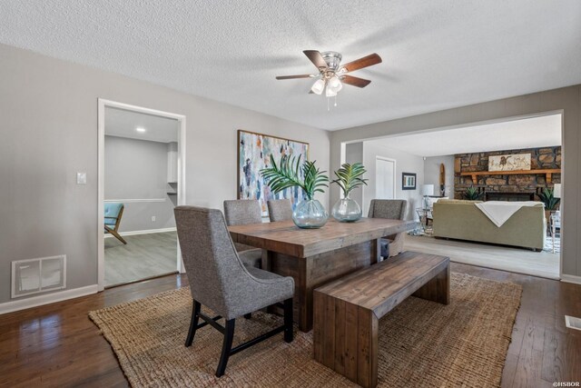 dining area featuring visible vents, dark wood-type flooring, ceiling fan, a stone fireplace, and a textured ceiling