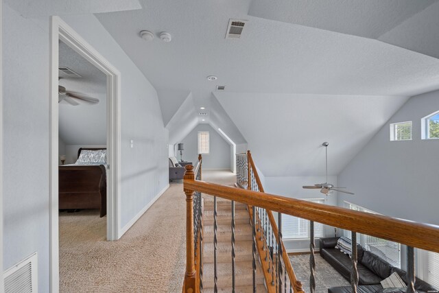 hallway featuring a textured ceiling, light colored carpet, an upstairs landing, visible vents, and vaulted ceiling