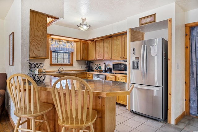 kitchen with tile counters, appliances with stainless steel finishes, brown cabinets, a peninsula, and a kitchen bar