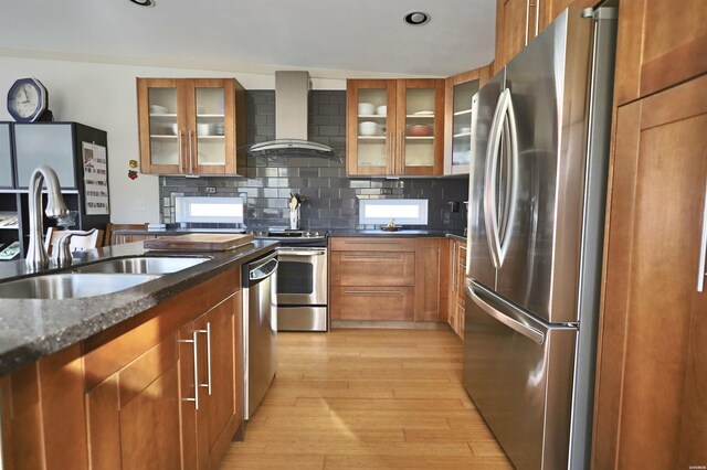 kitchen featuring brown cabinetry, wall chimney exhaust hood, glass insert cabinets, stainless steel appliances, and a sink