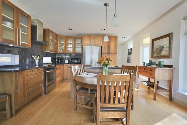 kitchen featuring wall chimney exhaust hood, glass insert cabinets, brown cabinets, appliances with stainless steel finishes, and pendant lighting