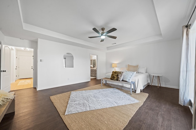 bedroom featuring dark wood-style floors, a tray ceiling, ensuite bathroom, ceiling fan, and baseboards