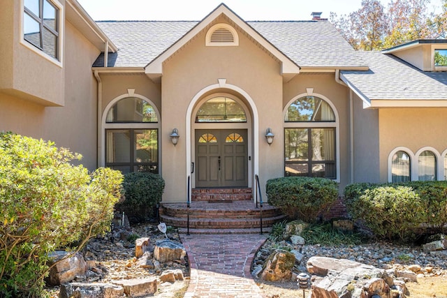 entrance to property with a shingled roof, visible vents, and stucco siding