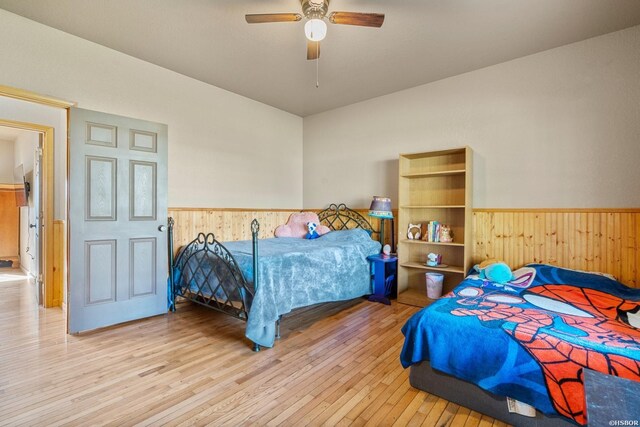 bedroom featuring light wood finished floors, wooden walls, a ceiling fan, and wainscoting