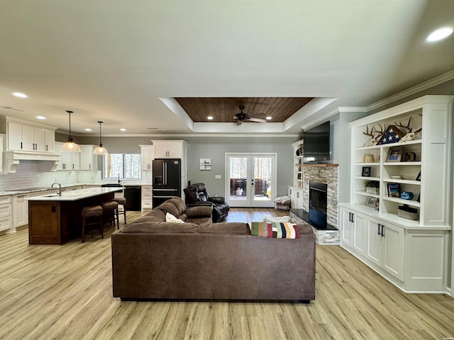 living area featuring a tray ceiling, a stone fireplace, recessed lighting, french doors, and light wood-style floors