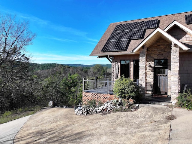 exterior space with stone siding, a shingled roof, roof mounted solar panels, and a wooded view